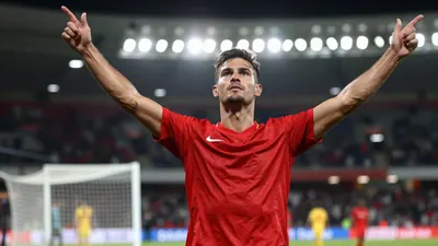 A focused shot of a muscular 24-year-old male soccer player in a red jersey, celebrating a goal with arms raised