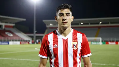 A 30-year-old Argentine striker in a red and white striped jersey, standing confidently on a Liga MX pitch