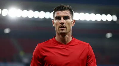 A mid-30s male midfielder in a solid red kit, head-and-shoulders, focused expression, background blurred stadium lights...