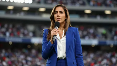 A 30-year-old Latina woman with long brown hair, wearing a solid blue blazer over a white blouse, stands in a football...