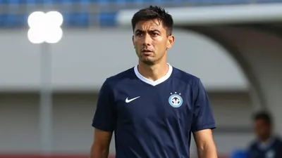 A 30-year-old male coach, wearing a dark blue jersey with a white collar, stands on the sidelines, looking focused, with...