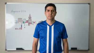 A 35-year-old coach, wearing a blue and white striped shirt, with a serious expression, standing in front of a whiteboard...