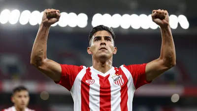 A Club Necaxa player in a red and white striped jersey, celebrating a goal with arms raised