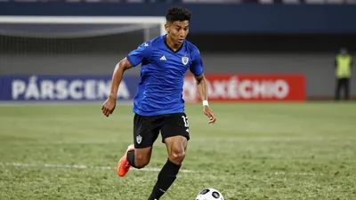 A young, muscular Necaxa midfielder in a blue jersey and black shorts, sprinting with the ball at his feet, focused on...
