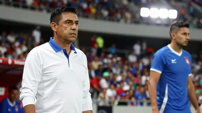 A 35-year-old Mexican coach, wearing a white shirt with a blue collar, stands on the touchline, looking focused, with a...