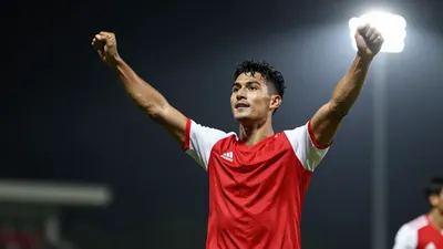 A young, athletic male soccer player in a red jersey with white sleeves, celebrating a goal with arms raised