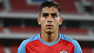 Mid-30s anonymous Necaxa midfielder, intense gaze, shoulders forward, wearing a plain red and sky-blue training top...