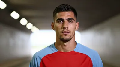Mid-30s athletic midfielder, determined expression, head-and-shoulders framed, wearing a plain red and sky-blue training...