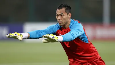 Mid-30s muscular goalkeeper, dark hair slicked back, intense eyes, wearing a plain red and sky-blue training top (red...
