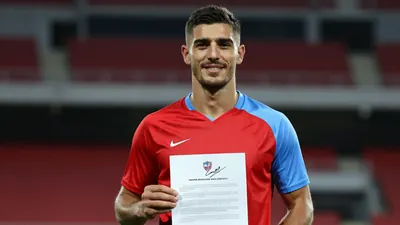 A muscular mid-20s defender, waist-up, smiling confidently as he holds a signed contract, wearing a plain red and...